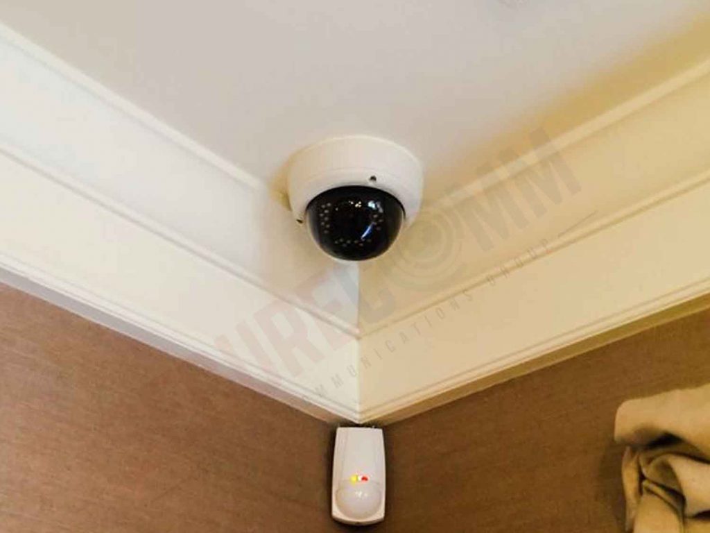 Indoor PTZ camera mounted on a ceiling with a motion detector on the wall, illustrating a typical surveillance setup.